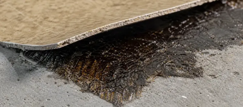 Angehobene Vinylbodenplatte mit sichtbarem schwarzem Teerklebstoff auf Betonuntergrund