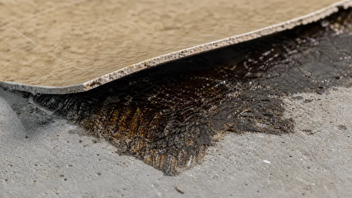 Angehobene Vinylbodenplatte mit sichtbarem schwarzem Teerklebstoff auf Betonuntergrund